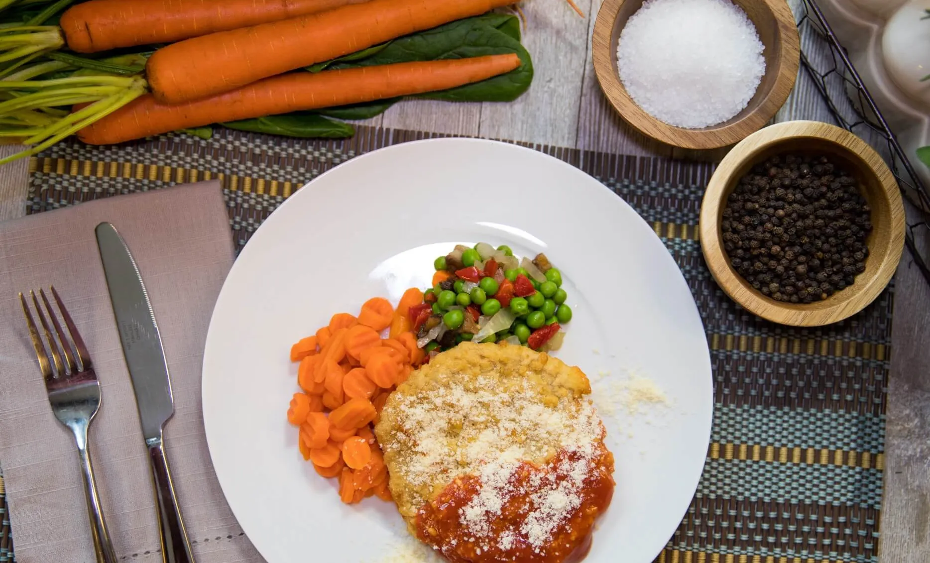 Chicken parmesan, carrots, and peas on white plate with whole carrots and spices in the background
