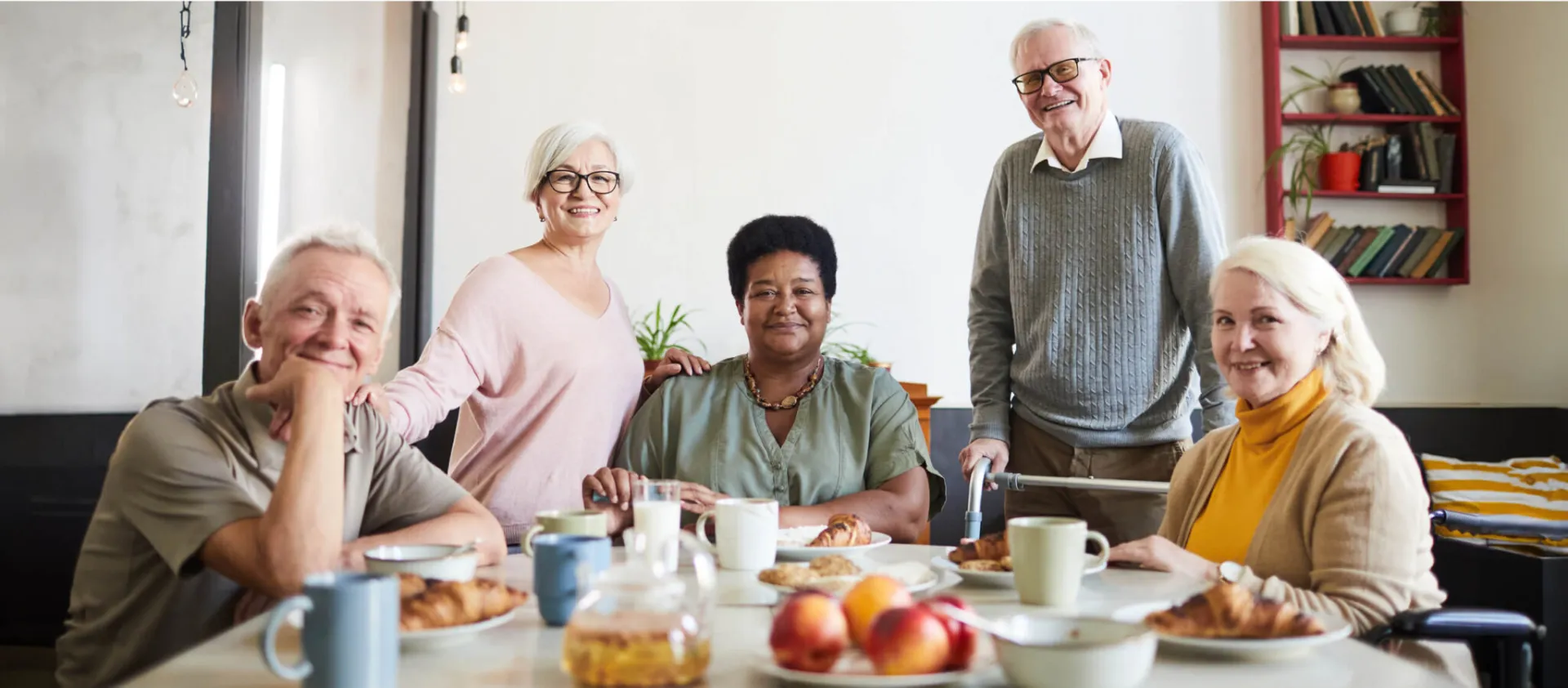 Five senior individuals happily gathered around a table with fruit, coffee and pastries