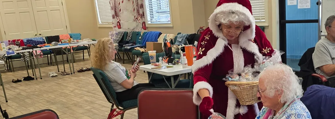Woman dressed as Mrs. Clause helping with a senior with holiday crafts at table