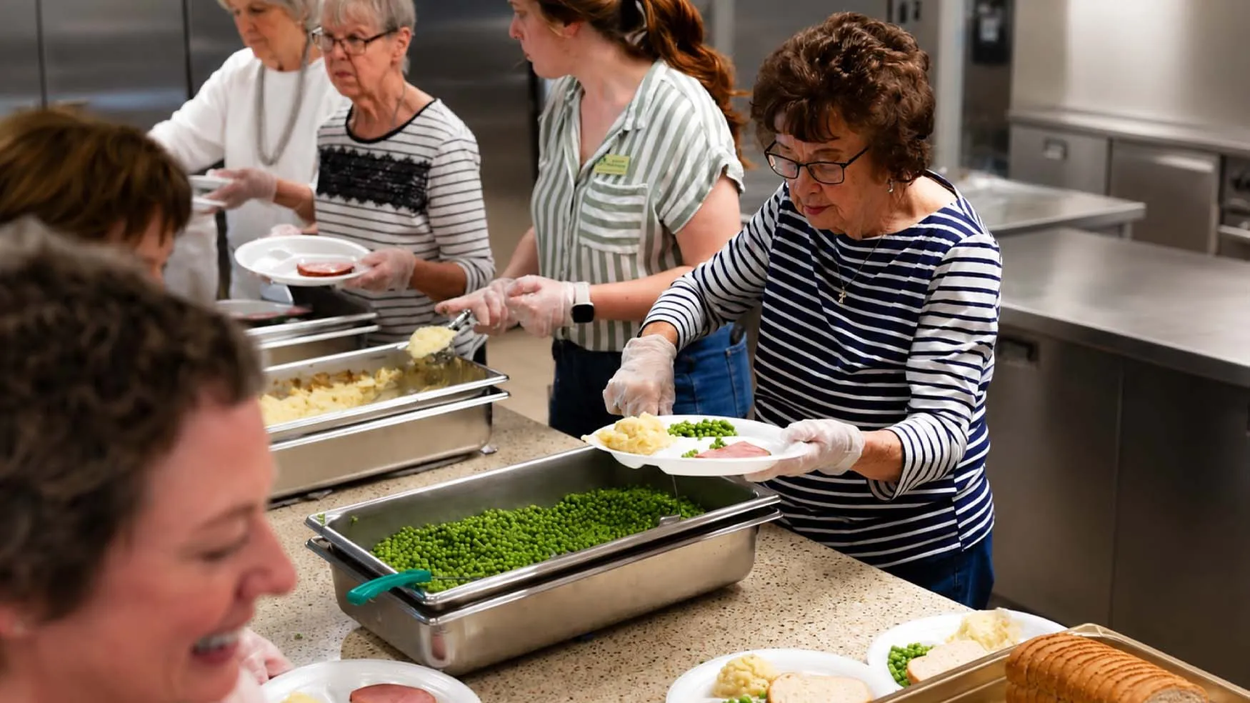 Volunteers serving a meal of ham, mashed potatoes, and peas to older adults in a community dining setting.
