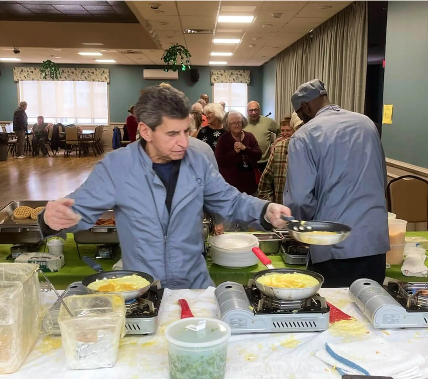 a senior man cooking eggs on portable burners in the middle of a gathering space