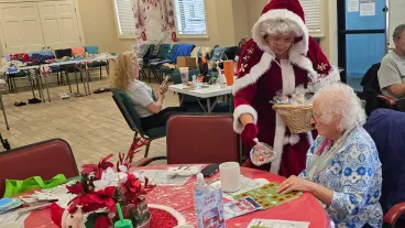 Woman dressed as Mrs. Clause helping with a senior with holiday crafts at table