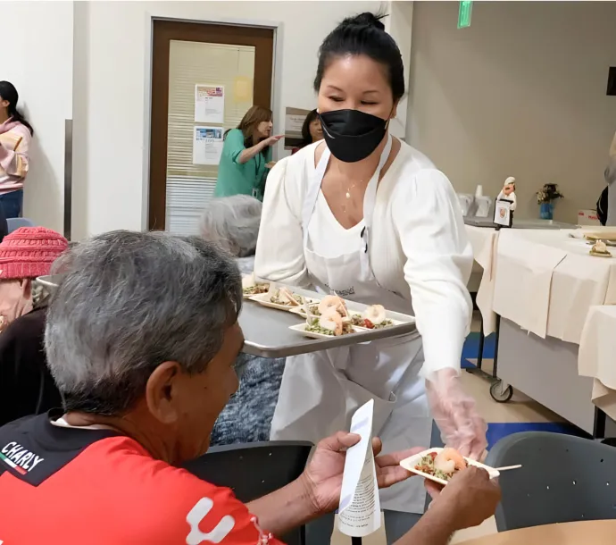 Employee wearing mask and apron, passing a shrimp dish to man at table